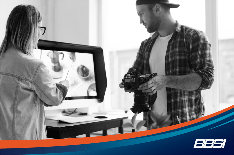 A man is holding a professional camera toward a product during a photo shoot for social media. A woman is next to him, directing the shoot.