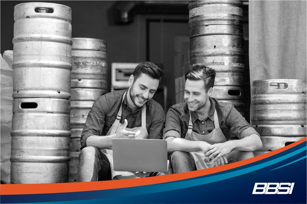 Two brewers looking at a laptop in a brewery next to barrels