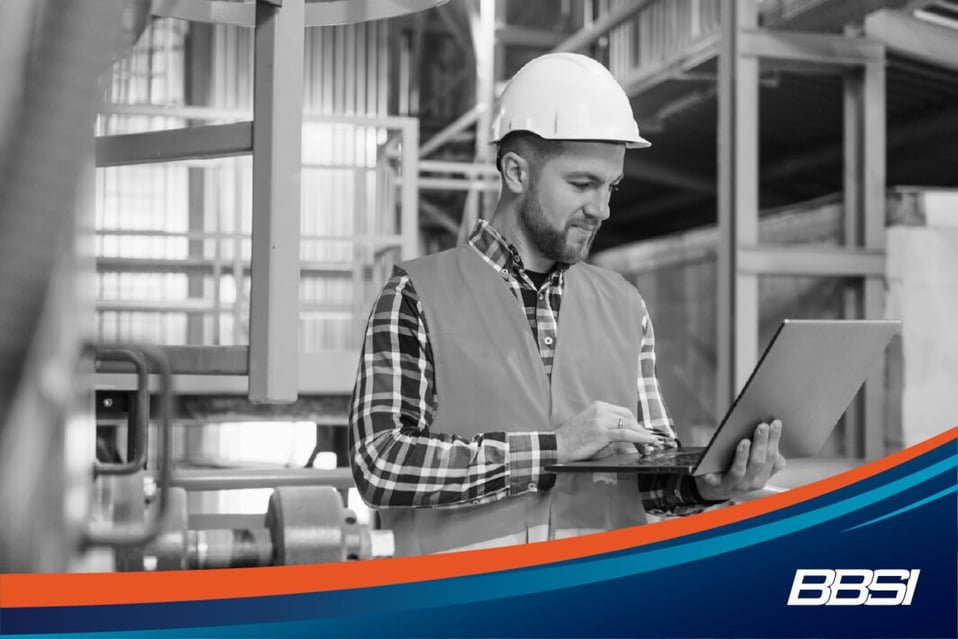 Construction worker wearing a neon orange safety vest and white hard hat in a warehouse looking at his laptop.