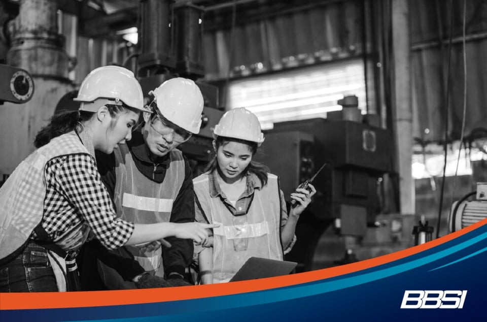 Three employees in PPE in a factory looking at a laptop and using a walkie talkie