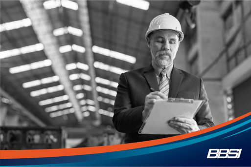 A factory owner, wearing a white hard hat and dark colored suit writing on a clipboard.