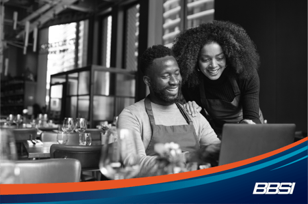 Two restaurant employees wearing aprons looking at a laptop screen sitting at a restaurant table.